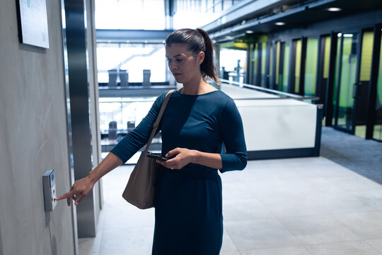 Businesswoman pressing elevator call button at office lobby, holding smartphone and bag, copy space