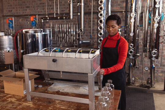 Young African American woman operating bottling machine on wooden table at distillery, copy space