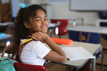 bi-racial schoolgirl sitting at desk smiling in classroom, holding pencil near orange pencil box
