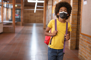 Boy standing in school hallway, with red backpack displaying purple straps and black face mask