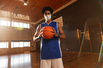 Man holding orange basketball on indoor gym court, wearing mask and blue wristbands