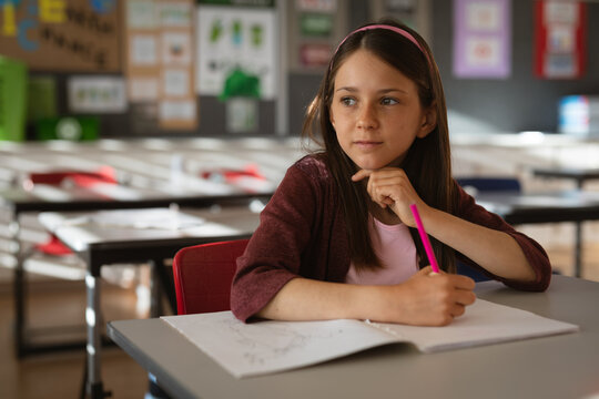 Elementary school girl writing in notebook at gray classroom desk in red chair with pink pencil