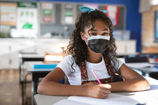 10-year-old girl sitting at desk in classroom writing in notebook with pencil wearing face mask