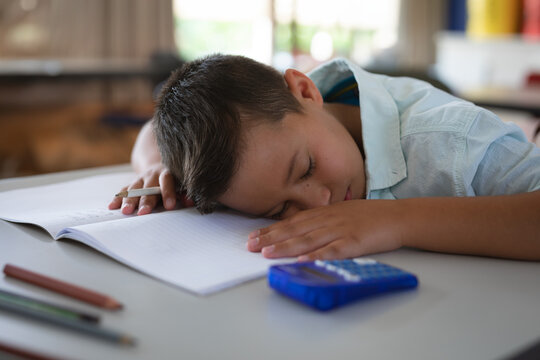 Eight year old boy sleeping on open notebook in classroom, with colored pencils, blue calculator