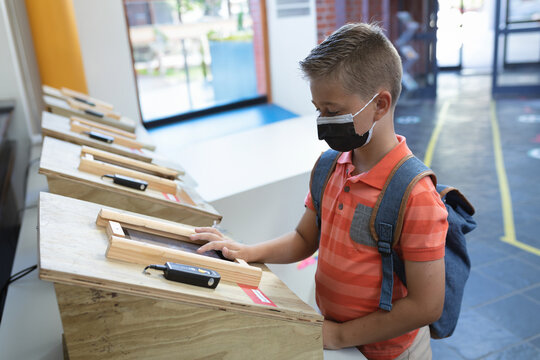 Boy wearing face mask and backpack touching device on wooden kiosk in school lobby, copy space