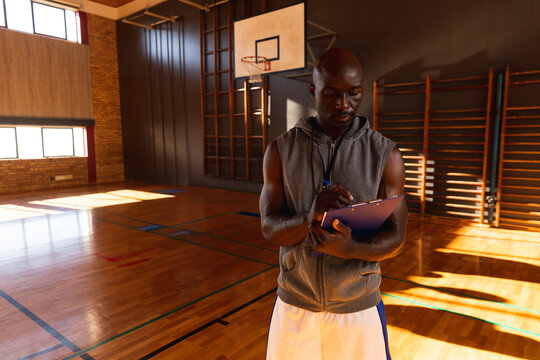 African American man coaching basketball drill by hoop in gym holding clipboard and pen, copy space