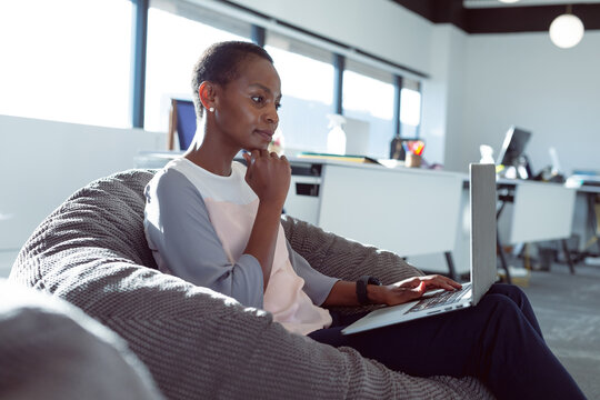 Mid adult African American woman typing on laptop sitting on gray beanbag chair in open-plan office