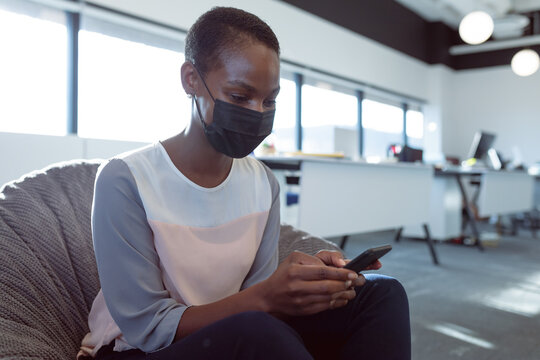 African American woman sitting on textured beanbag chair in open-plan office, viewing smartphone