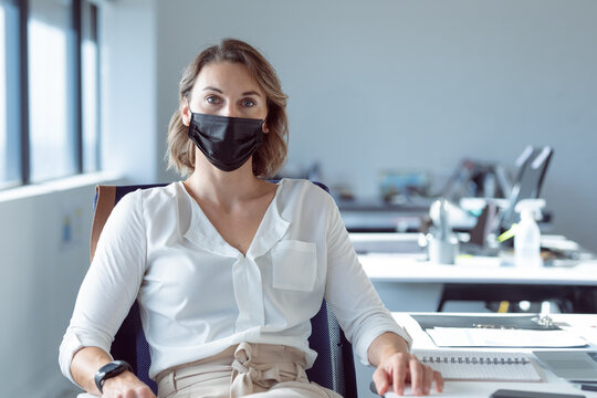Woman sitting at desk in modern open?plan office, writing in notebook while wearing black mask