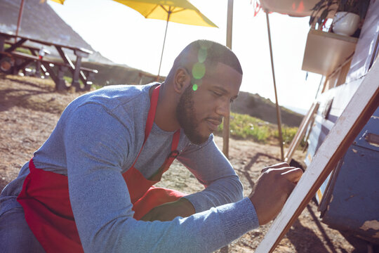 Young African American man kneeling writing on chalkboard menu by food trailer, wearing red apron