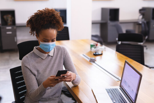 Woman wearing surgical mask scrolling smartphone at wooden conference table, viewing laptop graphs