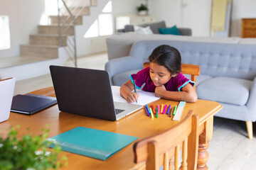 Girl child writing in notebook at wooden table in living room with colorful markers, copy space
