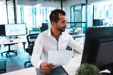 Man reviewing printed bar chart at modern open?plan office desk with computer monitor, potted plant