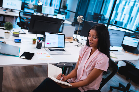 African American woman writing in notebook at desk in modern office with laptop, plant, copy space