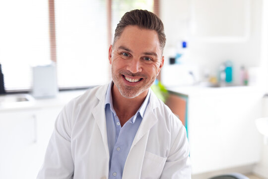 Male doctor smiling in exam room, wearing white lab coat under window blinds near sink