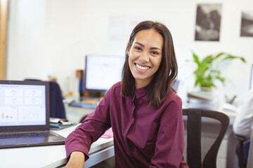 bi-racial woman leaning forward on desk in open-plan office displaying laptop charts while smiling