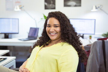 Mid-adult woman smiling at camera while working on laptop near potted plant in open-plan office