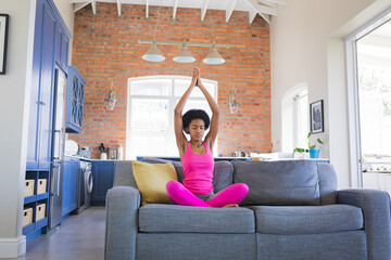 African American teenage girl practicing yoga on gray sofa in open-plan kitchen with blue cabinets