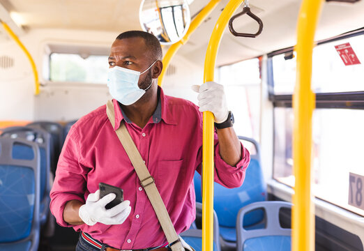 African American man standing in bus wearing face mask, holding smartphone and gripping yellow pole
