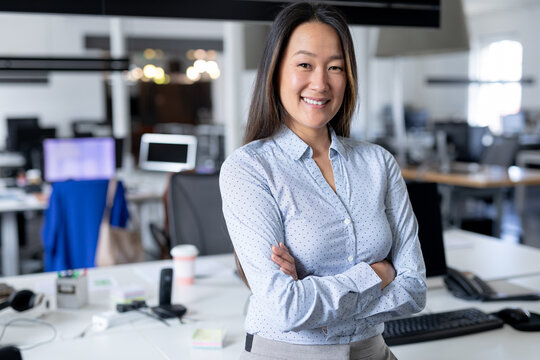 Mid adult Korean woman standing and smiling in modern office with computers, coffee cup, copy space