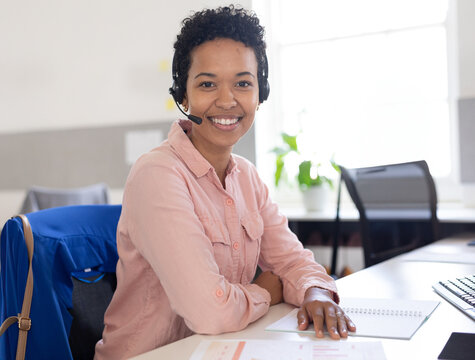 Woman sitting at modern office desk writing in notebook, with headset and scattered documents