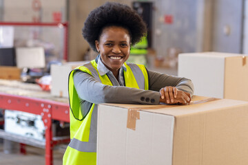 African American woman wearing neon vest leaning on cardboard box in warehouse, boosting logistics