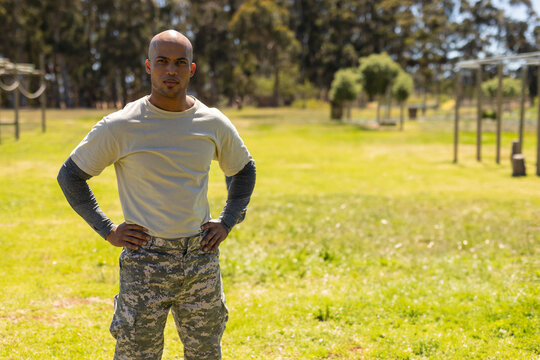 Man standing hands on hips in grassy training field, wearing camouflage pants and obstacle rings