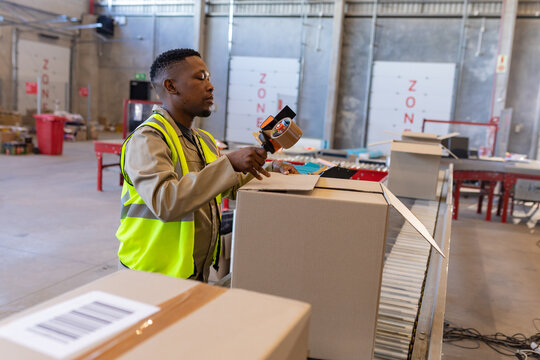 African American male warehouse worker sealing cardboard box at packing table, with tape dispenser