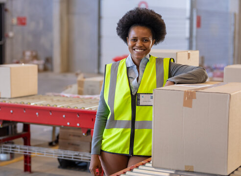Mid adult African American woman leaning on box at warehouse, wearing safety vest with ID badge