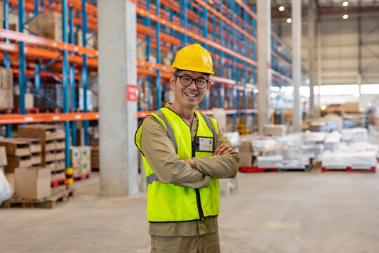 Asian man standing in warehouse near metal shelving racks, wearing safety vest and hard hat smiling