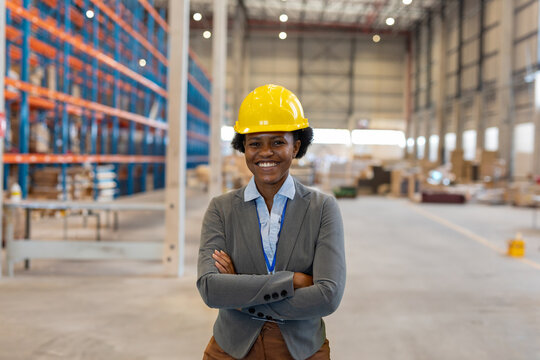 African American woman standing with crossed arms in large warehouse, wearing hardhat and ID badge