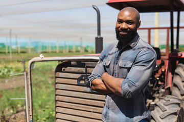 African American man leaning against red tractor under mesh netting at farm, crossing arms smiling
