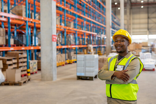 African American warehouse worker crossing arms in warehouse, wearing helmet and vest, copy space