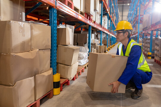 Mature Asian man crouching in warehouse aisle handling cardboard box, with shelving, copy space