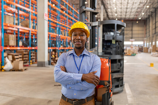 African American man standing with arms crossed in warehouse, wearing yellow hard hat and ID badge - Powered by Adobe