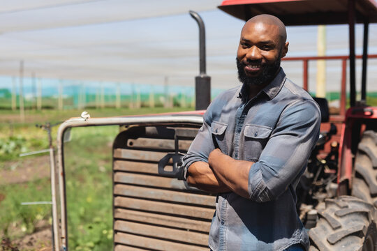 African American man leaning against red tractor under mesh netting at farm, crossing arms smiling - Powered by Adobe