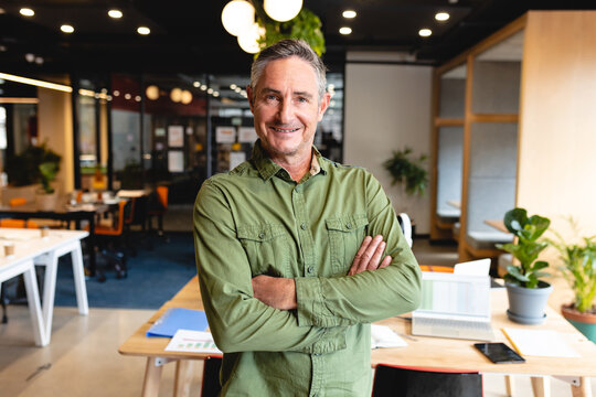 Mature adult man posing with arms crossed in modern open-plan office, showing laptop, potted plants