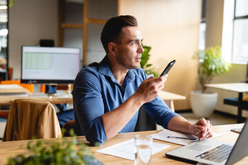 Young man speaking into smartphone at modern office desk, with laptop, papers and glass of water