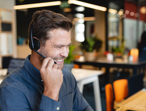 Mid-adult man speaking over headset at modern open-plan office desk, smiling brightly, copy space
