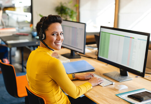 Man sitting at desk in open-plan office, wearing headset and using dual monitors for spreadsheets