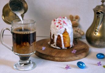 Easter cake covered with icing with a decor of a chocolate nest with eggs and a chicken topper on a white plate on a marble background. Easter table, treats, decor, traditions concept. Modern style, 