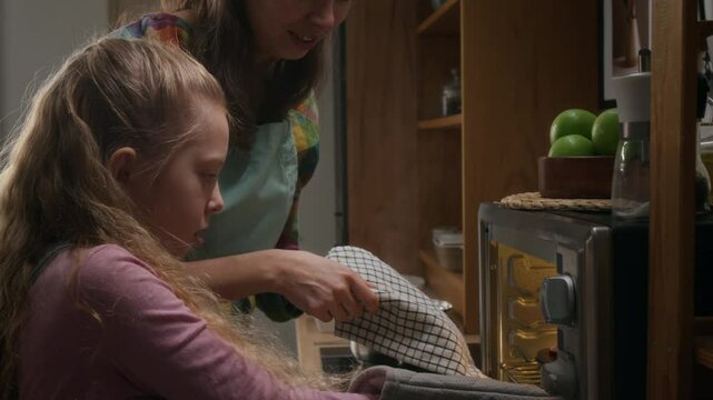 Medium arc shot of happy Caucasian woman helping daughter wearing padded glove to remove tray with freshly baked muffins out of hot oven, then smelling pastry aroma and smiling with joy