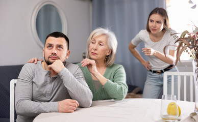 Offended man and his senior mother sitting at table at home, his wife standing behind and swearing at him.