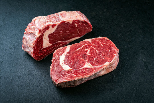 Dry aged raw beef entrecote cuts with fat marbling offered as close-up on a black rustic board