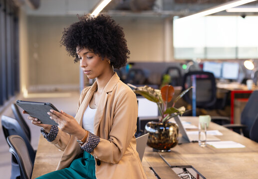 Young woman sitting on desk using tablet in open plan office, with laptop and plant