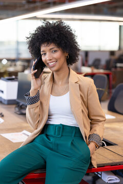 young adult woman sitting on wooden desk edge in modern office, talking on smartphone with papers