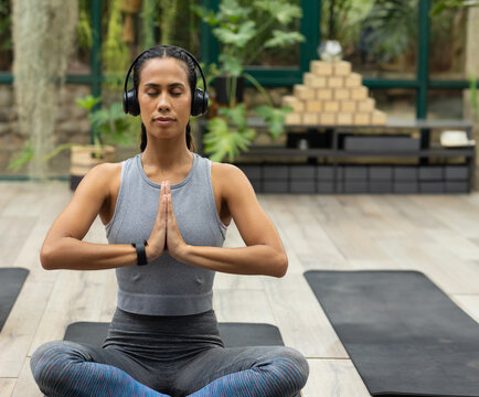 Woman meditating on yoga mat in studio with plants, wooden blocks, headphones and tracker