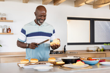 Senior African American man slicing bread roll at modern kitchen island, with knife and vegetables