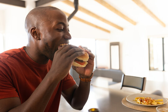 African American man eating hamburger at modern kitchen table, with plate of fries and wristwatch