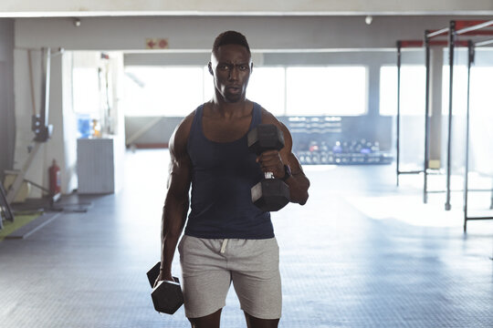 African American young man lifting heavy dumbbells in gym, with dumbbell rack and squat racks - Powered by Adobe